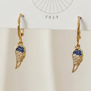 May include: Gold-coloured hoop earrings with a wing-shaped charm. The wing design incorporates blue and clear gemstones. The earrings are shown against a white background with the year 2023 printed on the top.