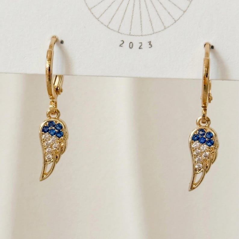 May include: Gold-coloured hoop earrings with a wing-shaped charm. The wing design incorporates blue and clear gemstones. The earrings are shown against a white background with the year 2023 printed on the top.