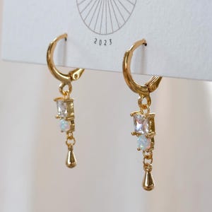 May include: A pair of gold hoop earrings featuring a dangling charm with a square clear stone, a round iridescent stone, and a small gold teardrop shape. The earrings are attached to a white display card with a sunburst logo and the text 2023.