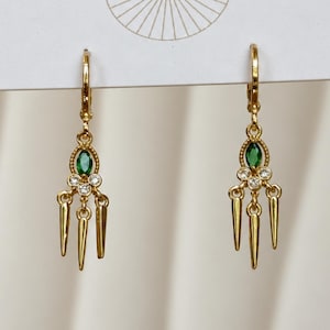 May include: A pair of gold-toned dangle earrings featuring a central oval-cut green gemstone, two small round clear stones, and three hanging gold spikes. The earrings are attached to a white display card with a thin gold line graphic at the top.