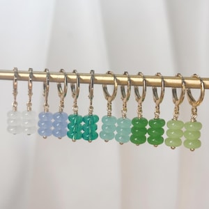 May include: A collection of twelve gold-tone hoop earrings arranged in a row, each featuring a dangling charm composed of three stacked, translucent, rounded beads in a gradient of colors including white, blue, teal, green, and lime.