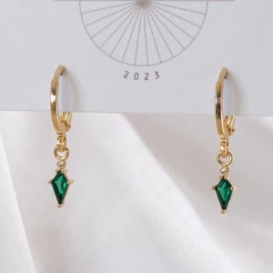 May include: A pair of gold hoop earrings featuring a small, kite-shaped green gemstone drop. Each earring has a tiny clear accent stone above the green gem. The earrings are attached to a white display card with a circular graphic and the text 2023.