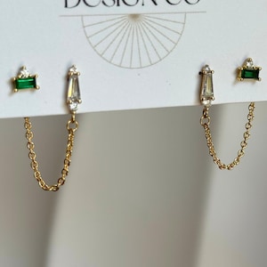 May include: A pair of gold chain earrings featuring two distinct studs each. One stud is a rectangular green stone with a small clear accent, and the other is a tapered baguette-shaped clear stone with a small clear accent, connected by a gold chain.