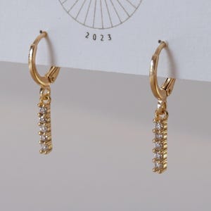 May include: A pair of gold hoop earrings with a hinged closure, each featuring a vertical drop bar pendant set with five small, clear, round-cut stones. The earrings are attached to a white display card printed with the year 2023.