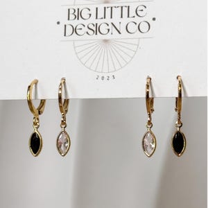 May include: Four gold-tone hoop earrings attached to a white display card labeled BIG LITTLE DESIGN CO 2023. The earrings feature dangling marquise-shaped charms, alternating between solid black and clear faceted stones in a symmetrical arrangement.