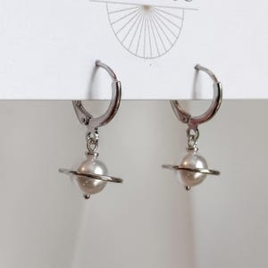 May include: A pair of silver-toned hoop earrings featuring a dangling charm shaped like a planet. Each charm consists of a white spherical bead encircled by a thin silver ring, suspended from a hinged hoop mechanism attached to a white display card.