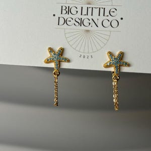 May include: A pair of gold-colored starfish earrings with turquoise and clear accents. Each earring has a dangling chain. The earrings are displayed on a white card with the text "JEWELRY & THINGS" and "BIG LITTLE DESIGN CO."