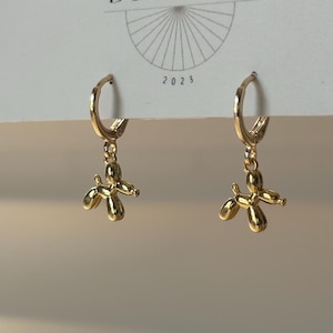May include: A pair of gold-colored hoop earrings. Each earring features a small, dangling charm shaped like a balloon dog. The earrings are displayed against a neutral background with the text "BIG LITTLE DESIGN CO" and the year "2023".