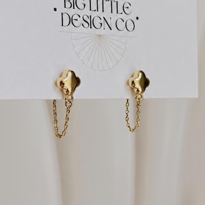 May include: A pair of gold-toned stud earrings featuring a four-leaf clover shape with a delicate chain drop attached to the base. The earrings are displayed on a white card printed with the text BIG LITTLE DESIGN CO and a sunburst graphic.