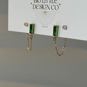 May include: Gold-toned earrings with emerald green rectangular stones and delicate chain accents. The earrings are displayed on a white card with the text "BIG LITTLE DESIGN CO" and the year "2023".