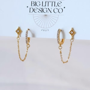 May include: A pair of gold-colored earrings with a starburst design and a chain. One earring is a hoop with small, clear stones. The other earring has a starburst stud and a chain that connects to the hoop. The earrings are on a white background with the text "BIG LITTLE DESIGN CO" and "2023".