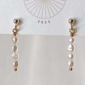 May include: A pair of gold-toned drop earrings featuring four small, irregular white pearls strung vertically on a thin wire, finished with a small gold bead at the bottom and a gold stud top. The earrings are attached to a white card with the text 2023.