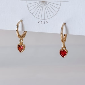May include: A pair of gold hoop earrings featuring small, dangling heart-shaped red gemstones. The earrings are attached to a white display card that features a thin line drawing of a sunburst design and the printed text 2023 at the center.