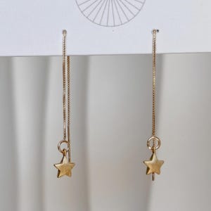 May include: A pair of gold-toned threader earrings featuring a delicate box chain design. Each earring terminates in a small, five-pointed star charm with a matte finish. The earrings are suspended from a white display card with a sunburst graphic.