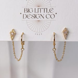 May include: A pair of gold-toned earrings on a white display card labeled BIG LITTLE DESIGN CO. Each earring features a diamond-shaped stud with small clear stones, connected by a delicate gold chain to a small hinged hoop earring component.