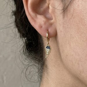 May include: A gold-coloured hoop earring with a wing-shaped charm. The charm features a cluster of small, sparkling white and dark blue stones. The earring is designed to be worn in the ear, a piece of jewellery.