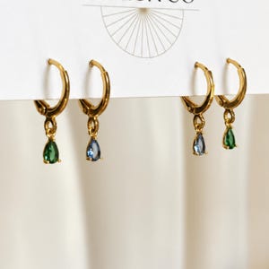 May include: Four gold hoop earrings attached to a white display card. Each hoop features a small, dangling teardrop-shaped gemstone. Two earrings have green teardrop stones, and two earrings have blue teardrop stones, all set in polished gold metal.