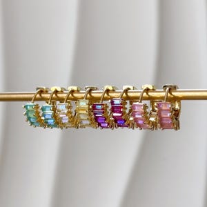 May include: A row of seven gold hoop earrings, each featuring a series of small, rectangular baguette-cut stones in a gradient of colors including teal, light blue, white, yellow, purple, magenta, and pink, all suspended on a thin gold rod.