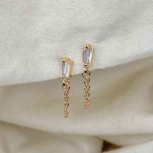 May include: A pair of gold earrings with a small, clear, rectangular stone and a delicate gold chain dangling from each.