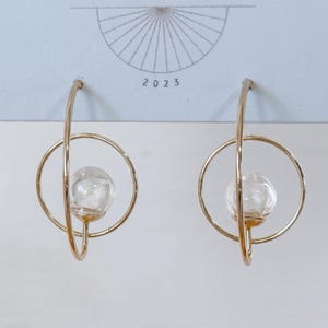 May include: A pair of gold-toned hoop earrings featuring a circular frame and a central clear glass sphere. The earrings are displayed on a light blue card with a sunburst graphic and the text 2023 printed in black at the top center.