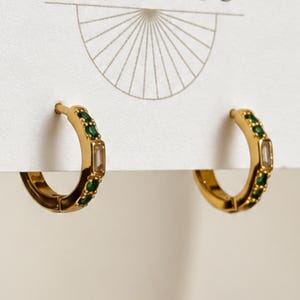 May include: A pair of gold-toned hoop earrings attached to a white display card. Each hoop features a series of small, round, vibrant green stones alternating with a single, clear, rectangular baguette-cut stone centered on the outer edge.