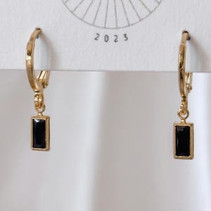 May include: A pair of gold-toned hoop earrings featuring a dangling rectangular black stone charm. The earrings are attached to a white display card with the text 2023 printed in black. The design includes a hinged closure mechanism for the hoops.