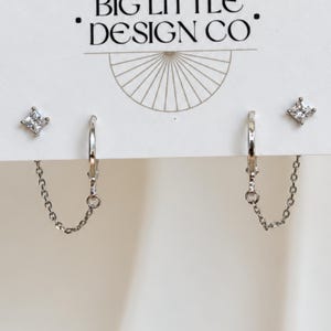 May include: Silver-toned earrings featuring a small hoop connected to a square-cut clear stone stud by a delicate chain. The display card is printed with the text BIG LITTLE DESIGN CO and a semi-circular line graphic above the jewelry pieces.