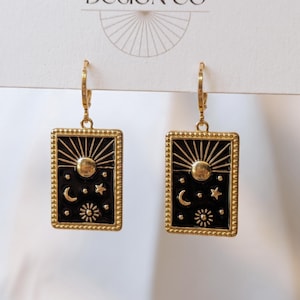 May include: A pair of gold-toned rectangular drop earrings featuring a black enamel inlay. The design depicts a celestial scene with a central sun, a crescent moon, and various stars. Each earring hangs from a gold-toned lever back closure.