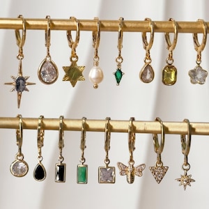 May include: A collection of sixteen gold-tone hoop earrings with various dangling charms, including a starburst, teardrop crystals, a pearl, a green kite shape, a yellow rectangle, a flower, a black teardrop, a bee, and a triangle.