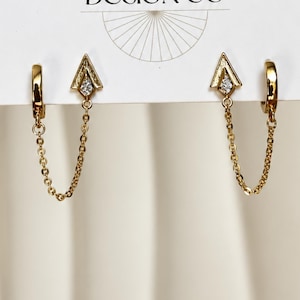 May include: A pair of gold-toned hoop earrings featuring a dangling chain that connects to a triangular charm. Each charm is accented with a small, clear, sparkling stone at its center. The earrings are displayed on a white card with a logo.