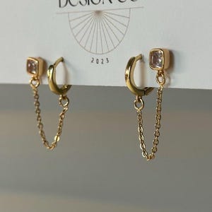 May include: Gold-toned earrings with a square, clear gemstone and a delicate chain. The earrings are attached to a gold hoop. The card behind the earrings has the text "BIG LITTLE DESIGN CO" and the year "2023".