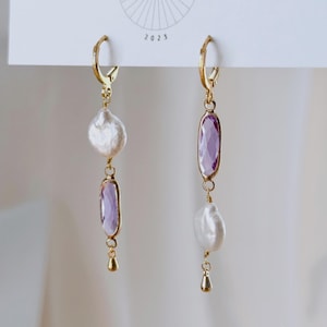 May include: A pair of gold-tone hoop earrings featuring asymmetrical drops. One earring displays a white baroque pearl above a faceted oval purple stone, while the other features a faceted oval purple stone above a white baroque pearl.