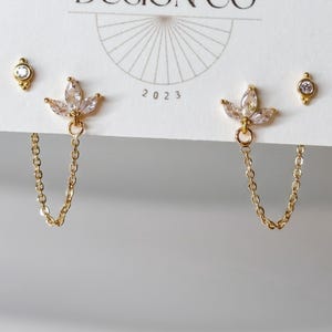 May include: Gold-tone stud earrings featuring a marquise-cut crystal cluster in a three-petal arrangement, connected by a delicate chain to a smaller round crystal stud. The jewelry is displayed on a white card with the text DESIGN CO 2023.