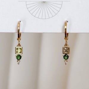 May include: A pair of gold hoop earrings featuring a vertical drop design. Each earring has a square-cut light green gemstone, a round dark green gemstone, and a small clear accent stone at the bottom, all set in a polished gold-tone metal.