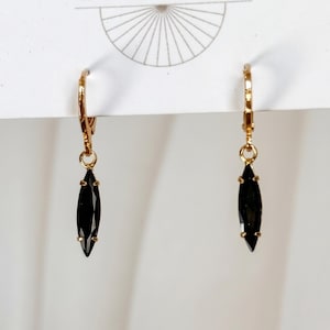 May include: A pair of gold-toned leverback earrings featuring elongated, marquise-cut black stones suspended from the hoops. The earrings are attached to a white display card that features a thin, circular line graphic at the top center.