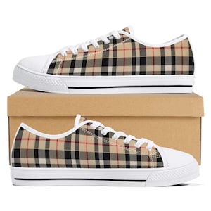 Puede incluir: Un par de zapatillas bajas con un patrón de cuadros en beige, negro y rojo. Los zapatos tienen cordones blancos, punteras de goma blanca y suelas de goma blanca con una fina raya negra a lo largo del lateral de la suela.