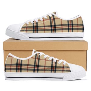 May include: A pair of low-top sneakers featuring a beige, black, and red plaid pattern. The shoes have white rubber toe caps, white laces, and white soles with a thin black stripe detail. The sneakers are positioned on top of a brown cardboard box.