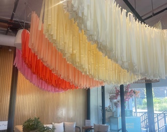 May include: A cafe interior featuring a decorative ceiling installation of cascading paper streamers in a gradient of colors, from pink to orange and yellow. The space includes seating, wooden planters with greenery, and a sign that reads "Natalie Blooms".