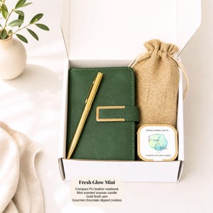 Pu&ograve; includere: Una confezione regalo con un taccuino in pelle verde, una penna dorata, una candela profumata alla cera di soia e un piccolo sacco di iuta. La confezione contiene anche biscotti al cioccolato. Il testo "Fresh Glow Mini" &egrave; visibile.