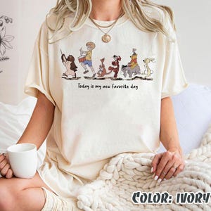 May include: Ivory-colored t-shirt featuring a cartoon illustration of characters in a line. The text "Today is my new favorite day" is printed below the characters. The shirt is a relaxed fit.