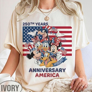 以下が含まれることがあります： アイボリー色のTシャツ。アメリカ国旗を背景に、ミッキーマウス、ミニーマウス、ドナルドダック、グーフィー、プルートなどのディズニーキャラクターが描かれています。「250th Years Anniversary America」の文字入り。