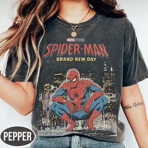 Może przedstawiać: Ciemnoszara koszulka z grafiką Spider-Mana w pozycji kucznej na tle panoramy miasta. Na koszulce widnieje napis "MARVEL STUDIOS SPIDER-MAN BRAND NEW DAY" w kolorze czerwonym i niebieskim.