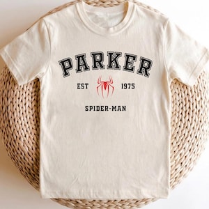 May include: Cream-colored t-shirt with the word "PARKER" in black arched lettering. Below the word "PARKER" is a red spider emblem, with the text "EST 1975" on either side. The words "SPIDER-MAN" are printed below the emblem.
