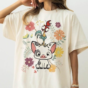 May include: Ivory-colored t-shirt featuring a cartoon pig with a rooster on its head, surrounded by colorful flowers and foliage. The text "COLOR: IVORY" is printed at the bottom. A casual, oversized fit.