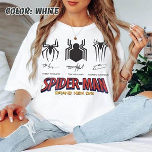 May include: White t-shirt featuring three black spider emblems and the text "SPIDER-MAN BRAND NEW DAY". The shirt also includes the names of Tobey Maguire, Tom Holland, and Andrew Garfield.