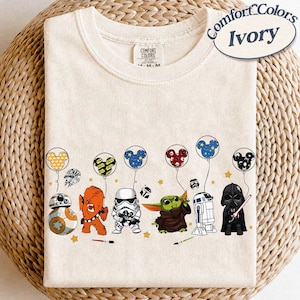 May include: Ivory-colored Comfort Colors t-shirt featuring Star Wars characters holding Mickey Mouse-shaped balloons. Characters include BB-8, Chewbacca, a Stormtrooper, Grogu, R2-D2, and Darth Vader. The shirt has a crew neck.