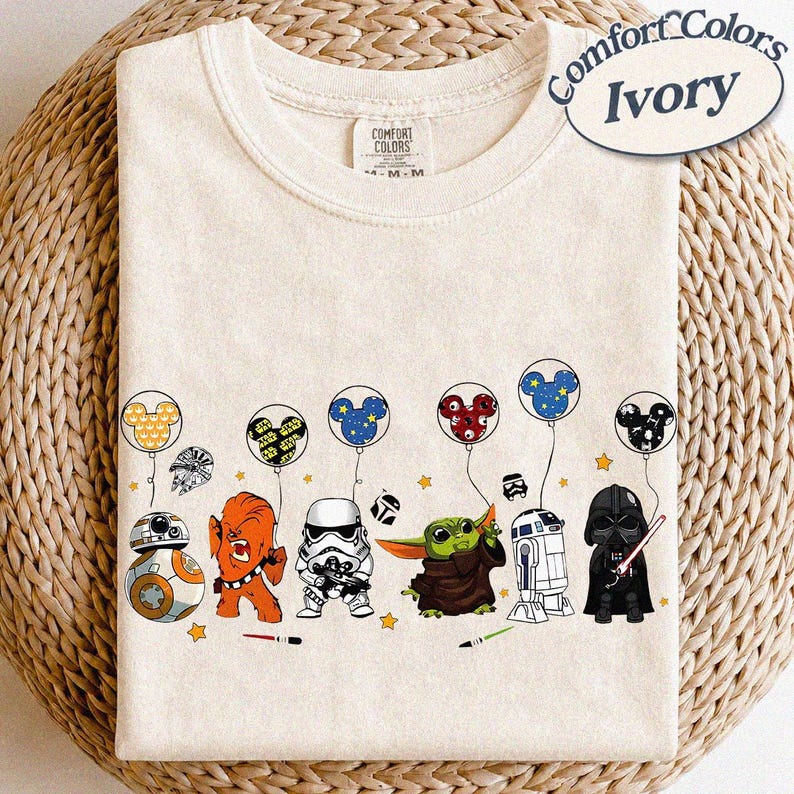 May include: Ivory-colored Comfort Colors t-shirt featuring Star Wars characters holding Mickey Mouse-shaped balloons. Characters include BB-8, Chewbacca, a Stormtrooper, Grogu, R2-D2, and Darth Vader. The shirt has a crew neck.