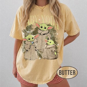 May include: A yellow short-sleeve t-shirt featuring a graphic of three green creatures in tan robes with the text BABY YODA printed in large light yellow letters above them. The design includes white lightning bolt accents and small stars.