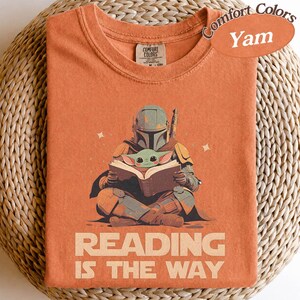 May include: Orange Comfort Colors t-shirt featuring a graphic of the Mandalorian reading to Grogu. The text "READING IS THE WAY" is printed below the illustration. The shirt has a crew neck and short sleeves.