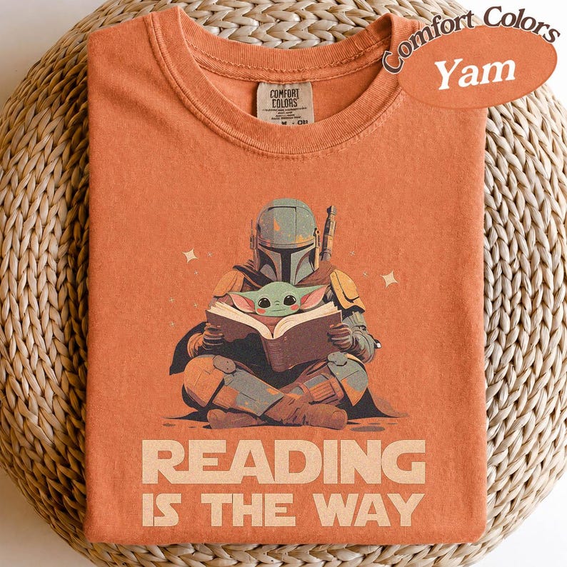 May include: Orange Comfort Colors t-shirt featuring a graphic of the Mandalorian reading to Grogu. The text "READING IS THE WAY" is printed below the illustration. The shirt has a crew neck and short sleeves.